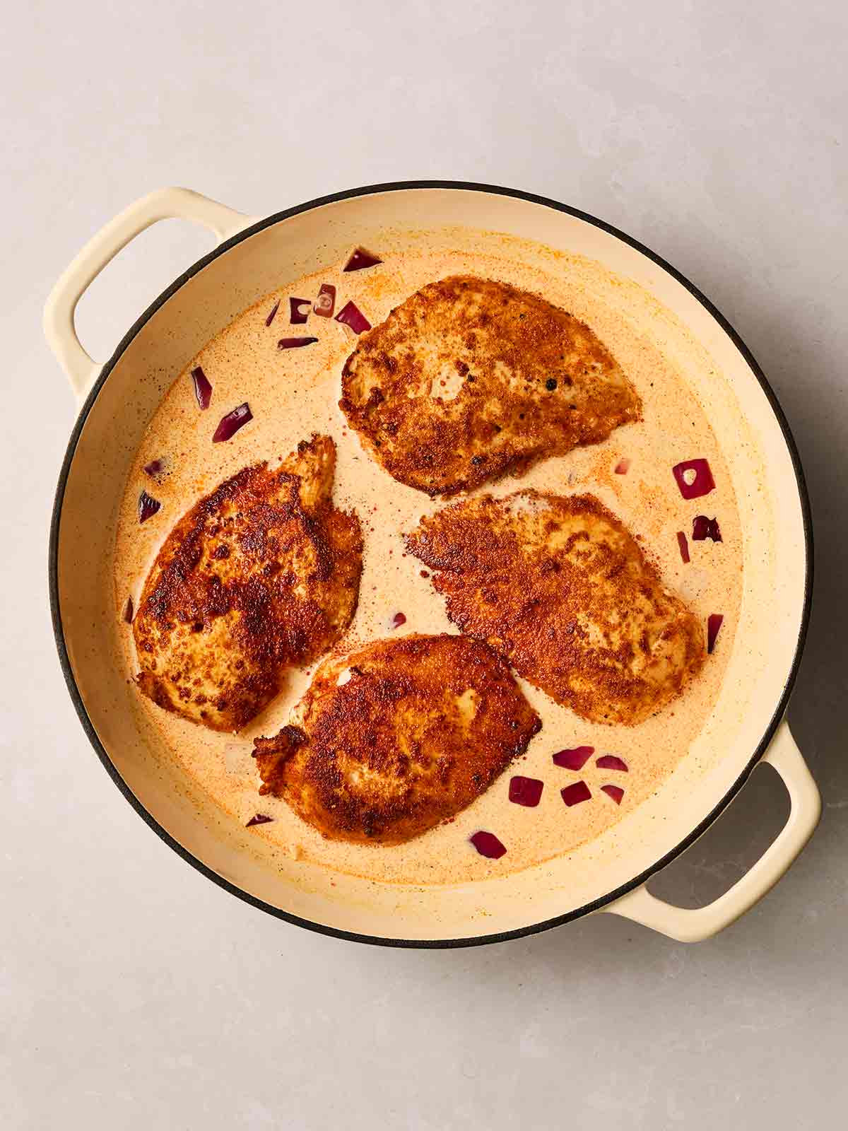 A pan filled with four cooked chicken breast with a red paprika coating, in a creamy sauce with red onions.