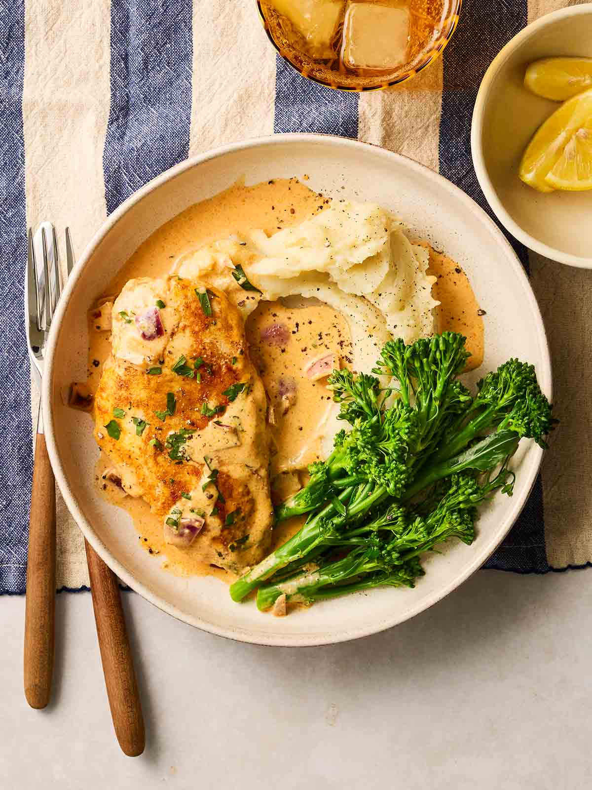 A plate on a table with a blue and white tablecloth, filled with paprika chicken in a creamy sauce, served with mash potato and broccoli.