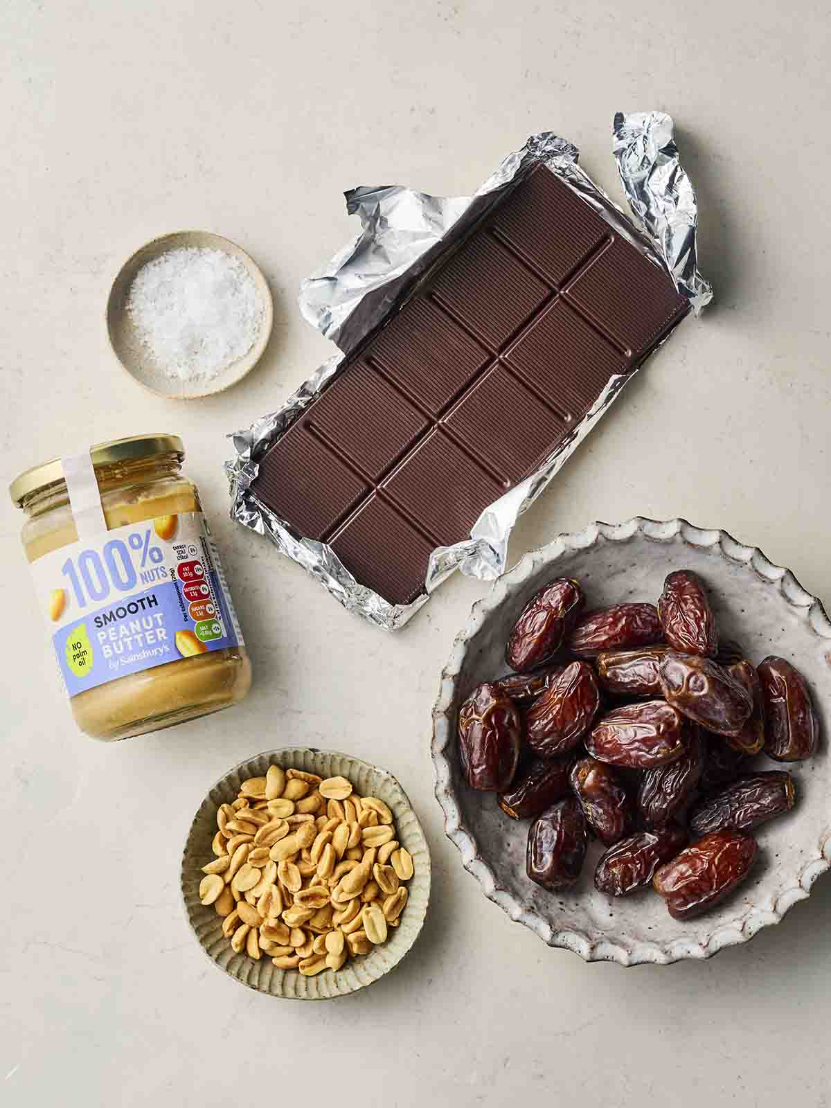 The ingredients for making Date Bark laid out on a white counter, including medjool dates, chocolate, peanut butter, salt and peanuts.