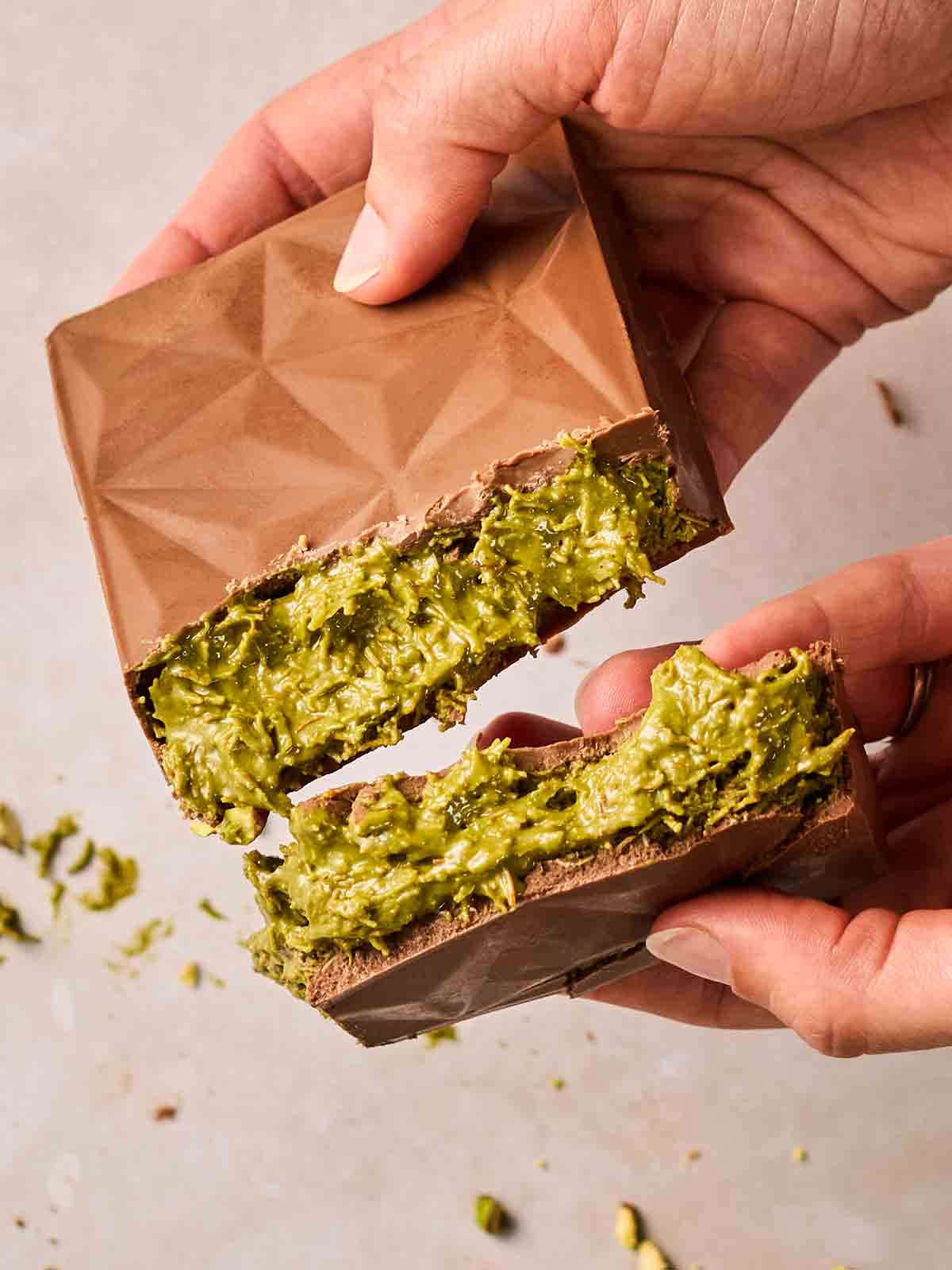 Two hands holding two pieces of Dubai Chocolate that has been broken in half to show the green filling.