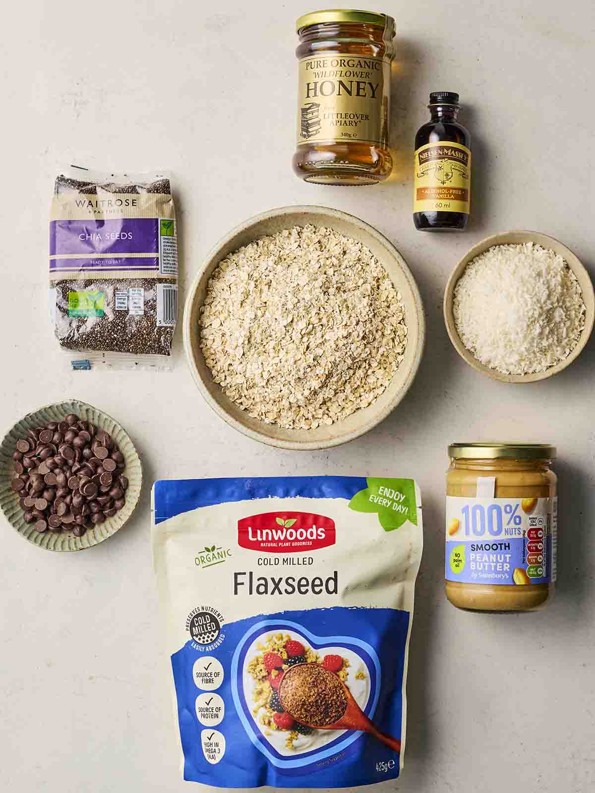 A collection of ingredients on a white surface, including oats, flaxseed, honey, dessicated coconut, peanut butter, chocolate chips, chia seeds and vanilla.