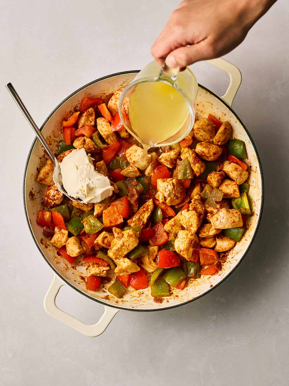 A pan of chicken fajita mix, with a hand pouring in stock and a spoon going in with cream cheese.