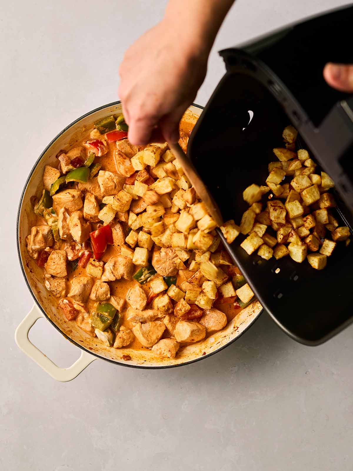 A pan of chicken and peppers in a sauce, with cubed potatoes being poured in from an air fryer pan with two hands.