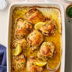 A baked lemon chicken dish with slices of lemons and cooked chicken thighs with herbs over the top.