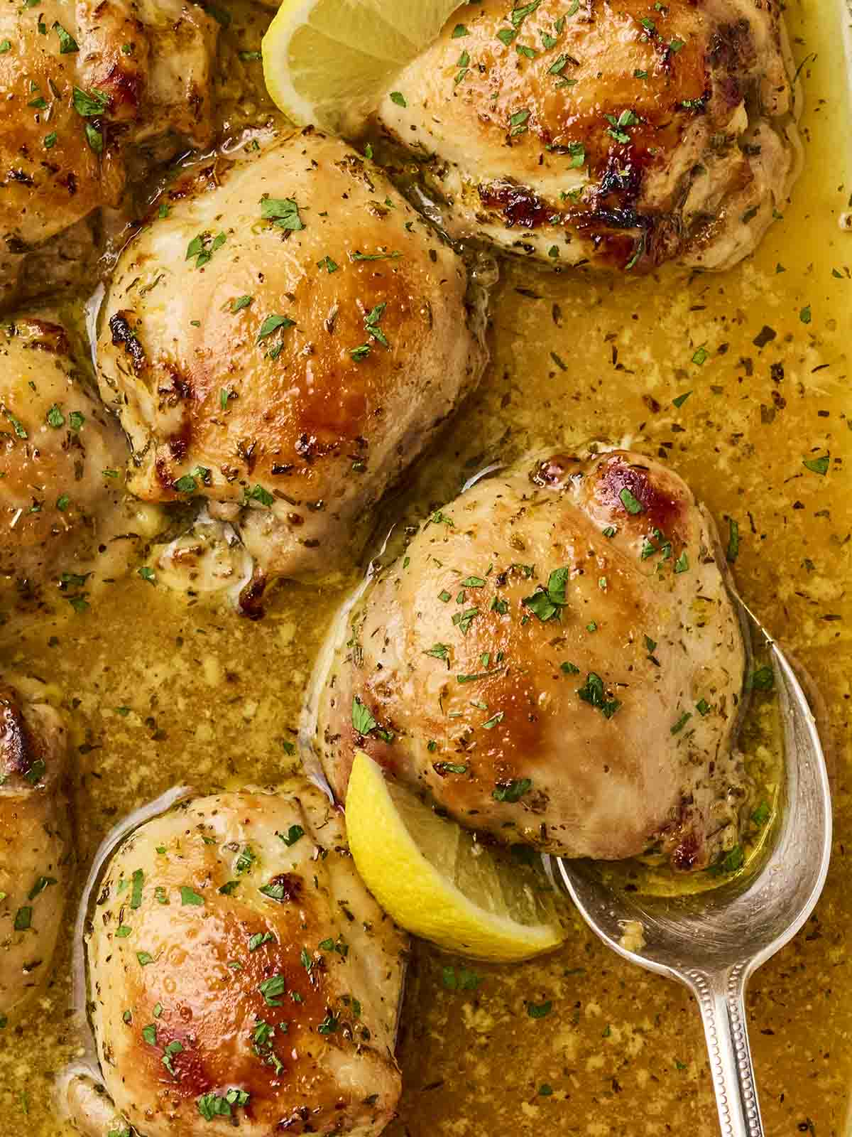 A close up of baked chicken thighs, soaked in lemon sauce and herbs, with a golden finish, with one being served in a spoon.