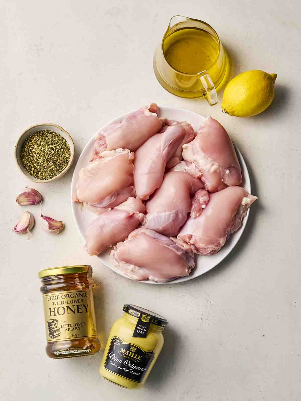Ingredients laid out on a counter top, including raw chicken thighs, honey, oil, lemon, garlic, oregano and mustard.