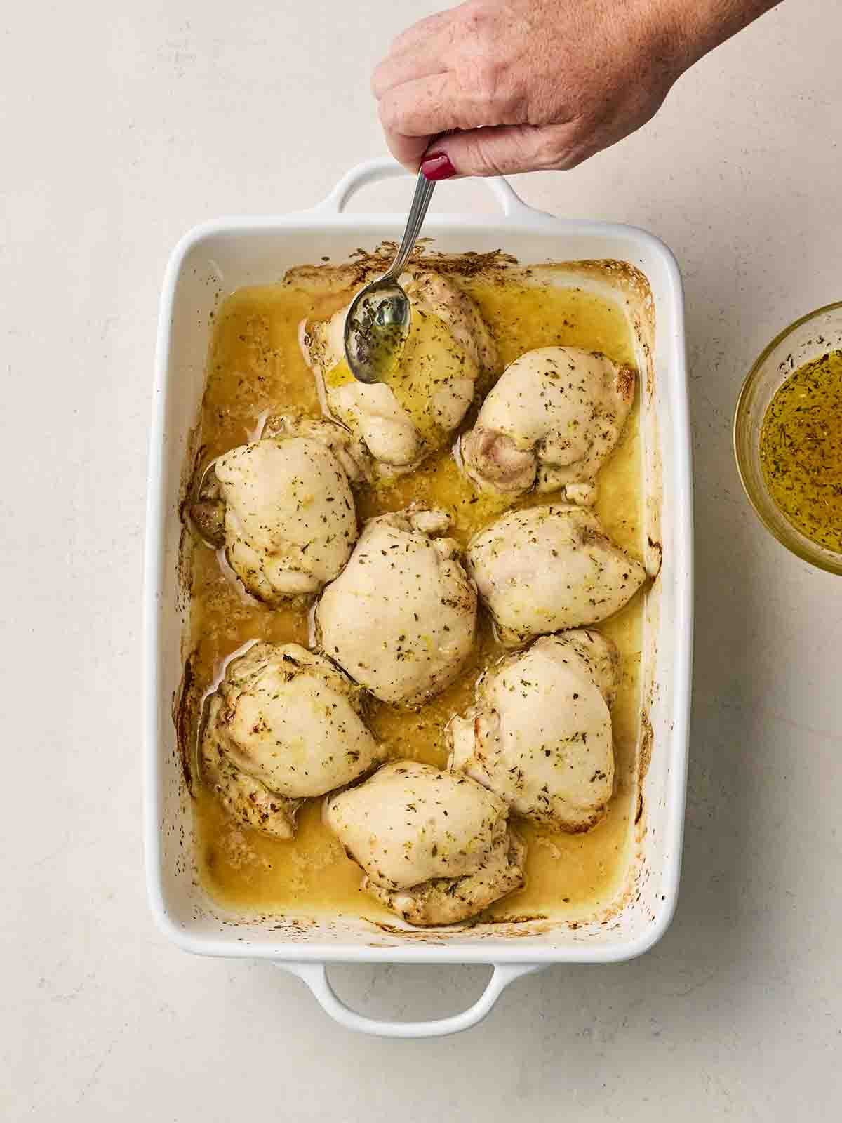 An oven dish with part-cooked chicken thighs, in a lemony sauce with oregano, with a hand spooning over the juices.