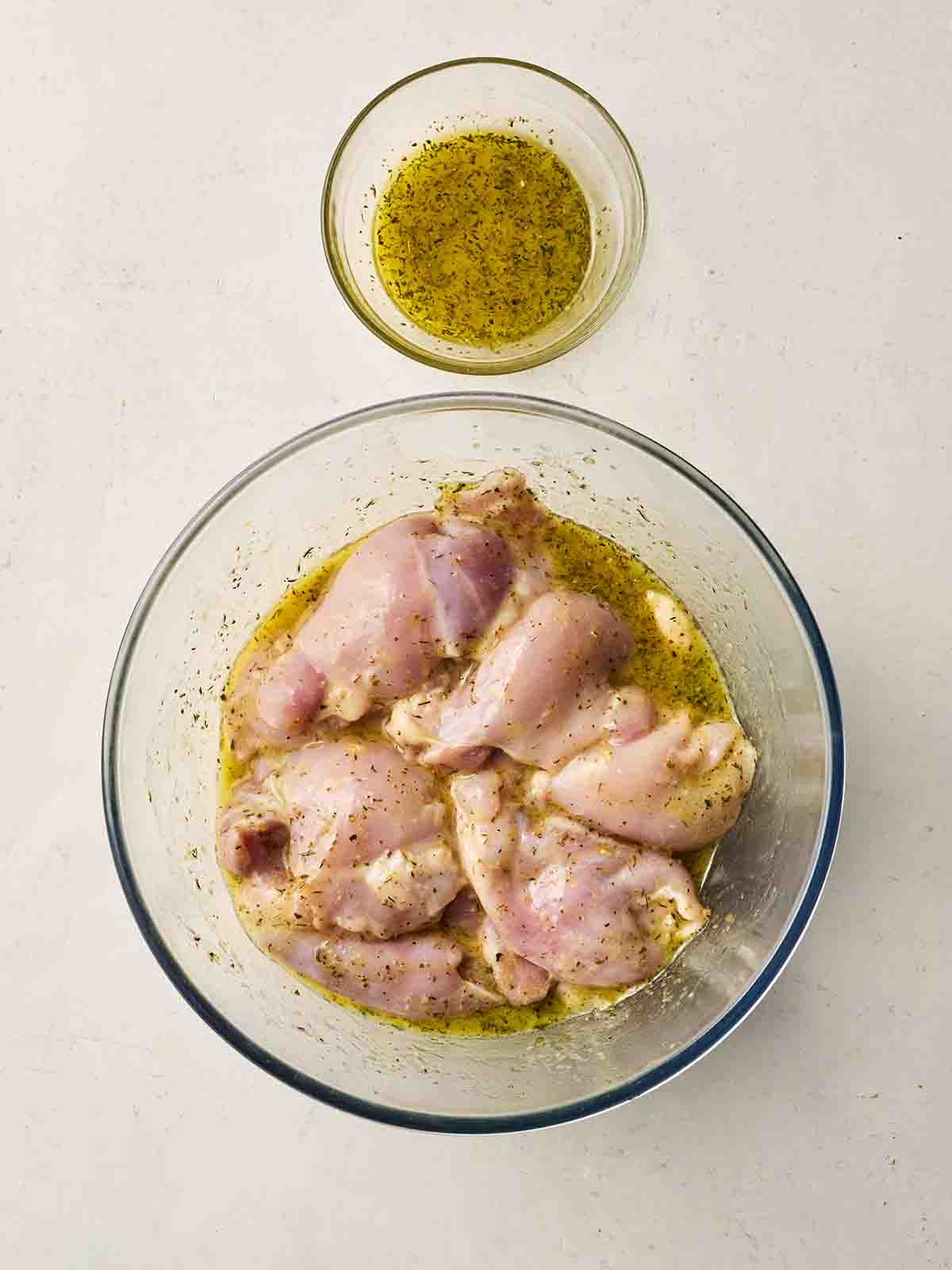 A glass bowl with marinading chicken in a lemony sauce, with the sauce in a bowl to the side.