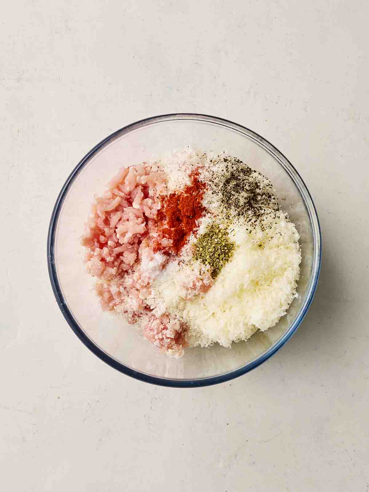 Raw ingredients in a glass bowl together, unmixed. Including parmesan, chicken mince, herbs and spices.