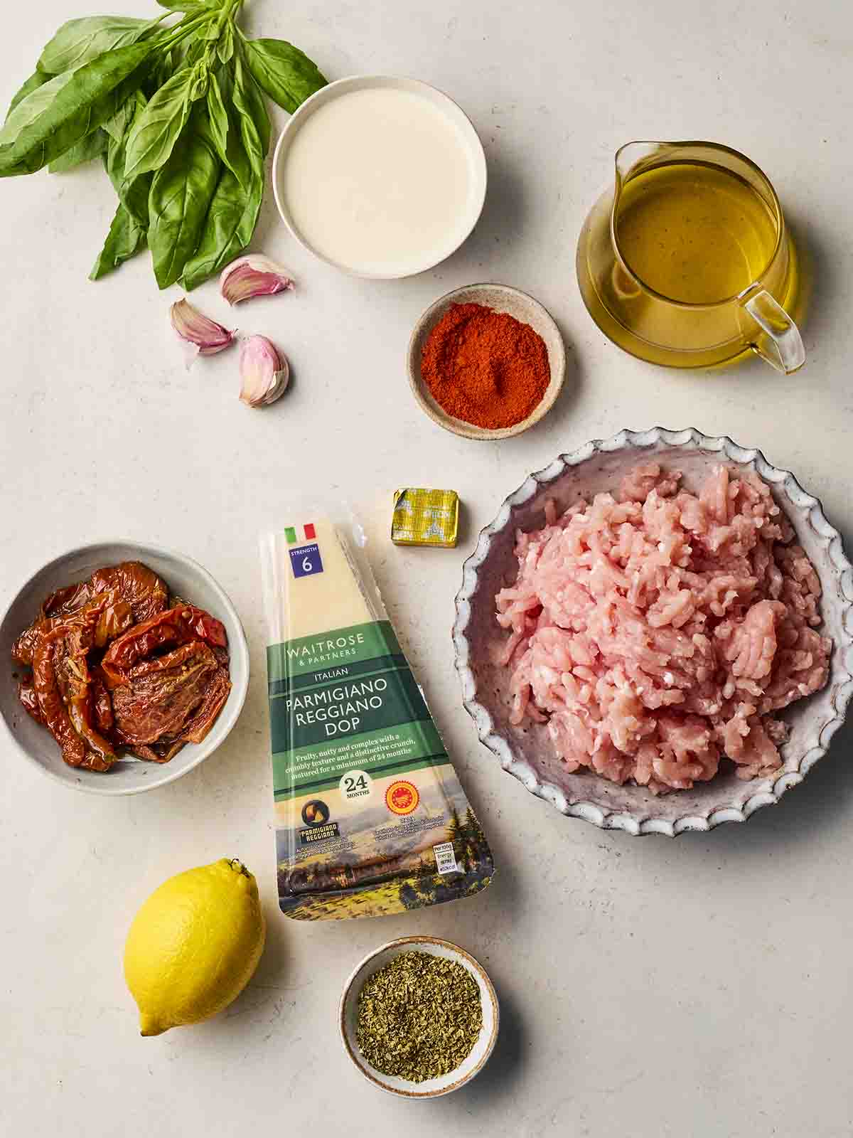 Raw ingredients laid out on a white counter top, including chicken mince, oil, herbs and spices, lemon, sun-dried tomatoes and basil.