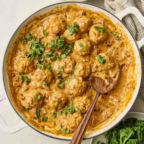 A large pan filled with creamy chicken meatballs for Marry Me Meatballs recipe, on table with a serving spoon and a bowl of mash and broccoli to serve.