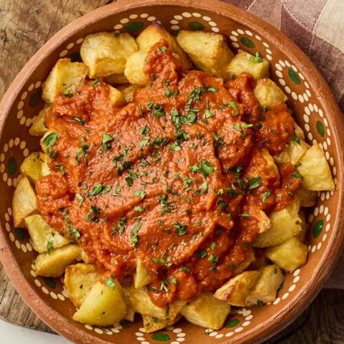 A bowl of Patatas Bravas tapas on a board with a tea towel.