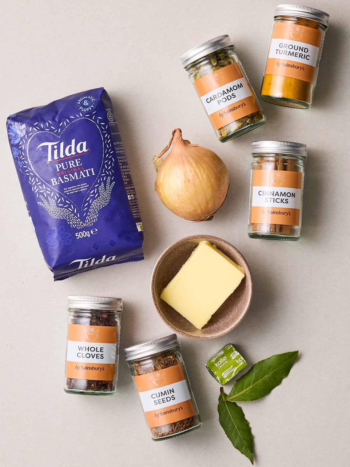 Ingredients laid out on a table for making Pilau Rice, including lots of spices, basmati rice, butter, onion, vegetable stock and bay leaves.