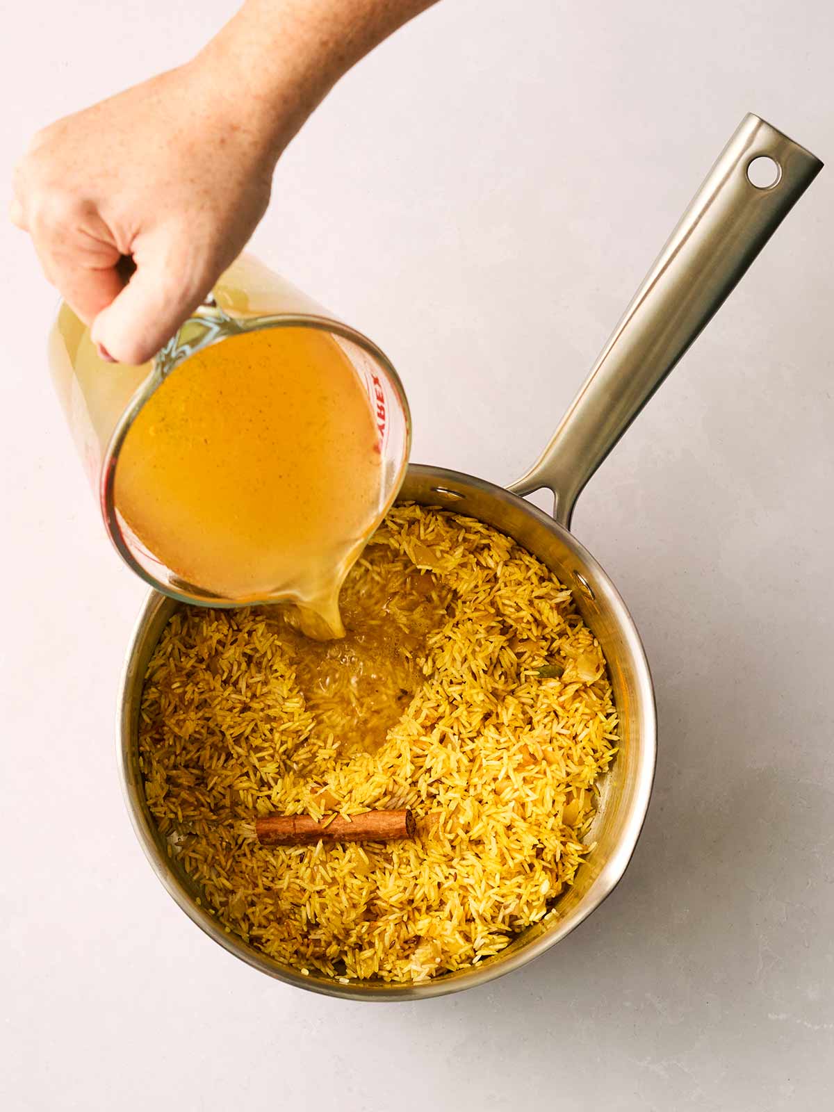 A pan of yellow rice with spices and hot stock being poured in.