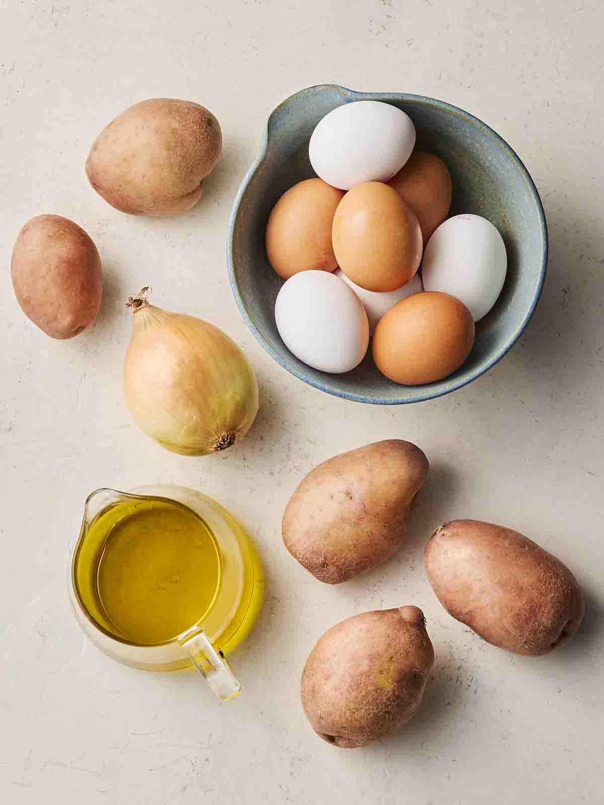 Ingredients laid out on a white counter, including potatoes, eggs, an onion and oil.