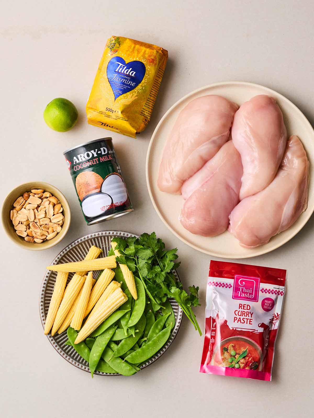 Raw ingredients laid out on a white surface, including raw chicken breast, veg and rice, Thai curry paste and coconut milk.