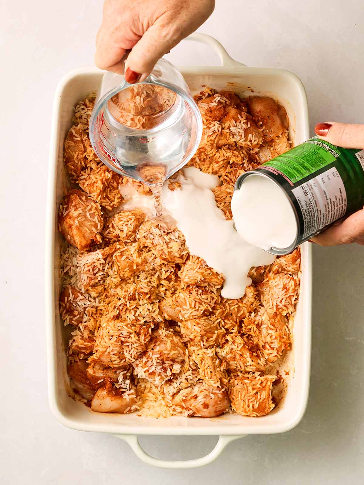 An oven dish with rice and chicken in, with two hands, one pouring coconut milk and one pouring a jug of water.