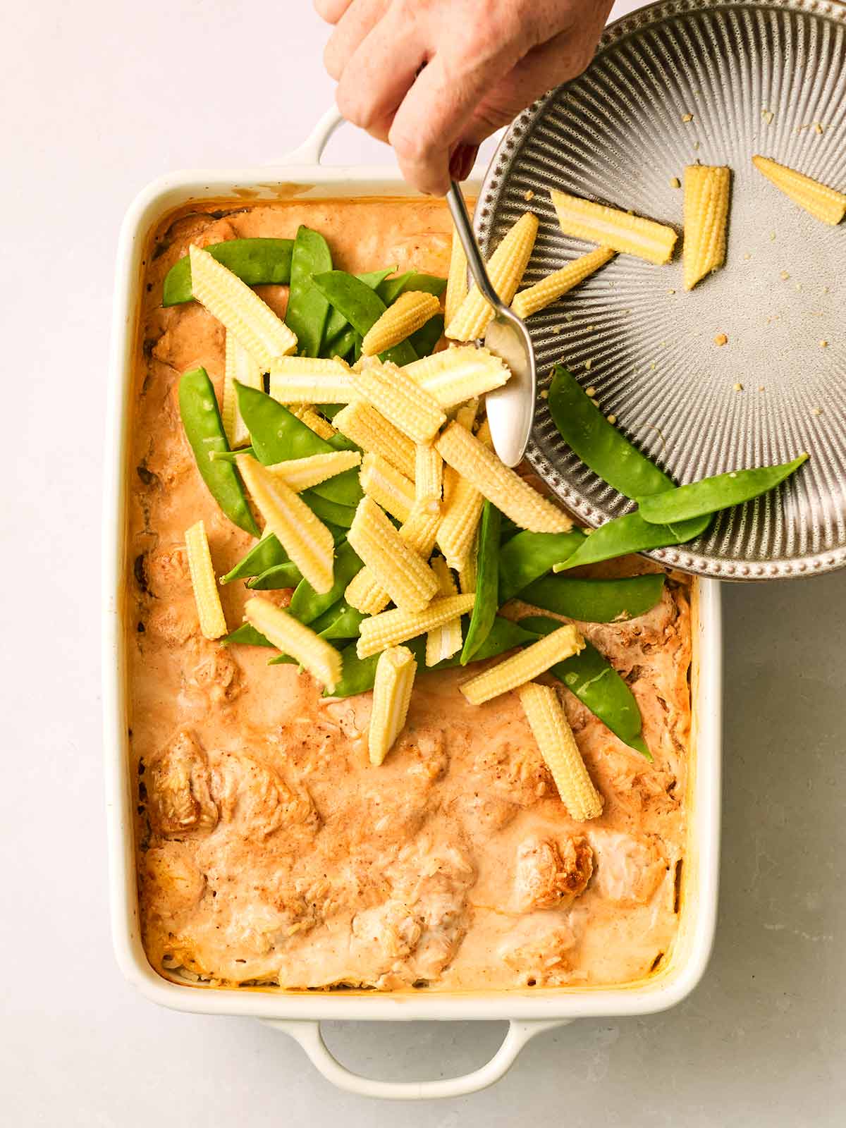 Vegetables being poured into an oven dish with chicken and rice in a curry sauce.