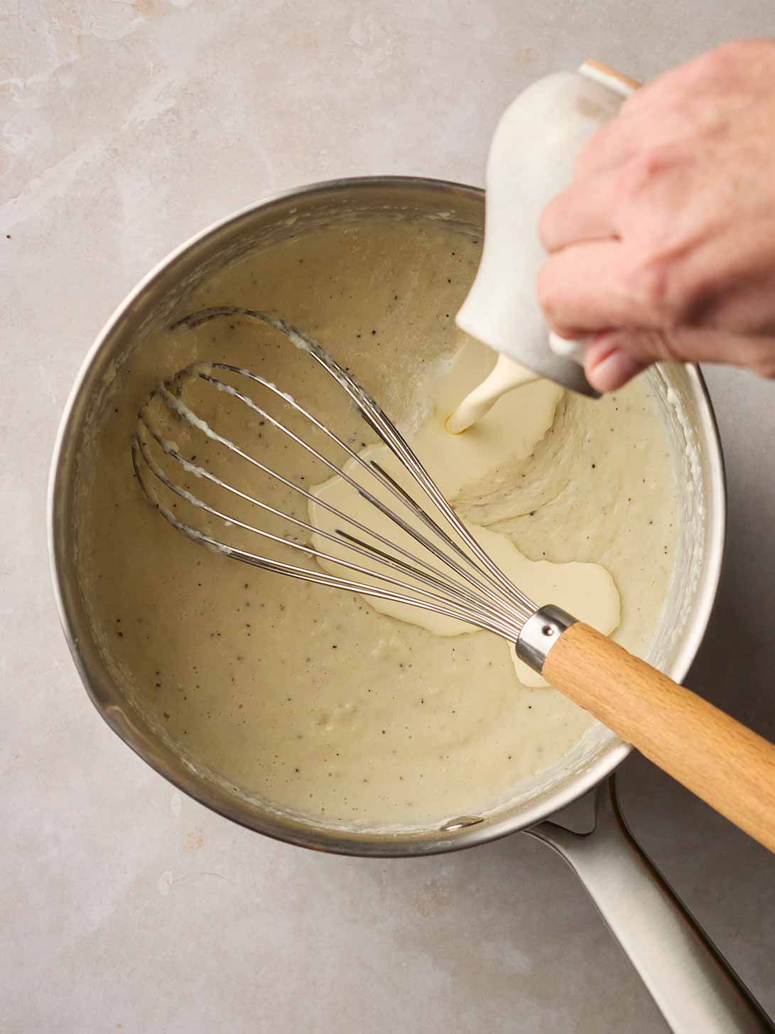 A hand pouring a jug of cream into a pan with a whisk for step 4 in the recipe for making Bread Sauce.