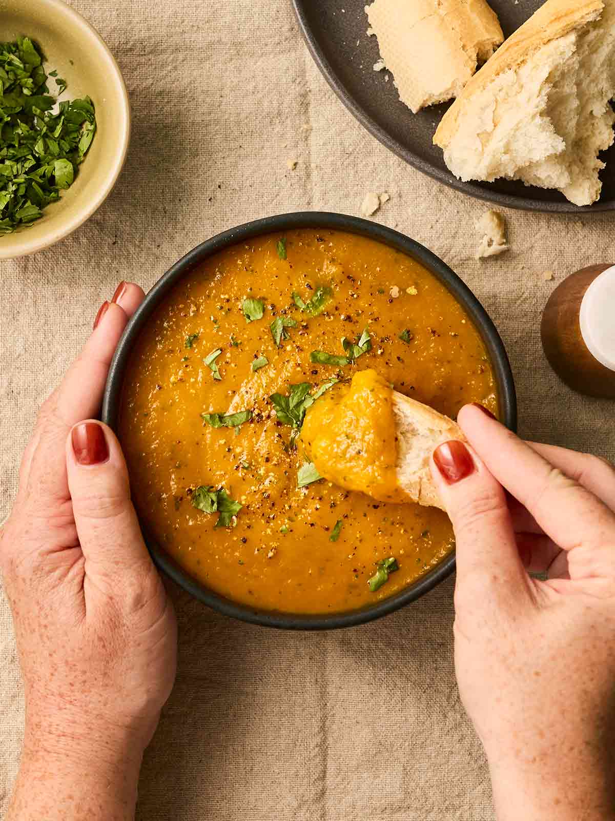 Two hands cradling a bowl of orange soup, with one hand dipping a piece of bread in.