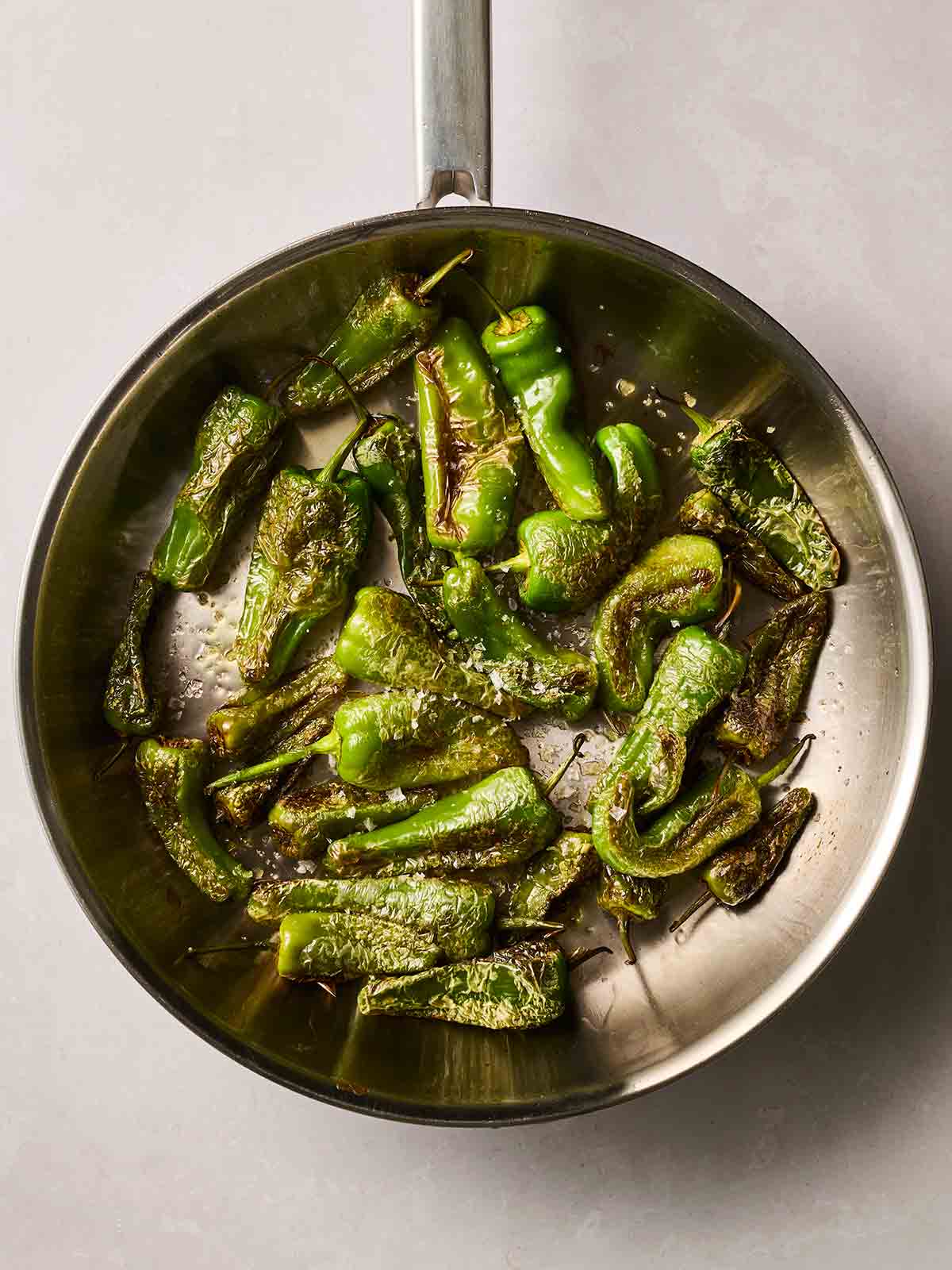 A pan filled with panfried padron peppers on a white surface.
