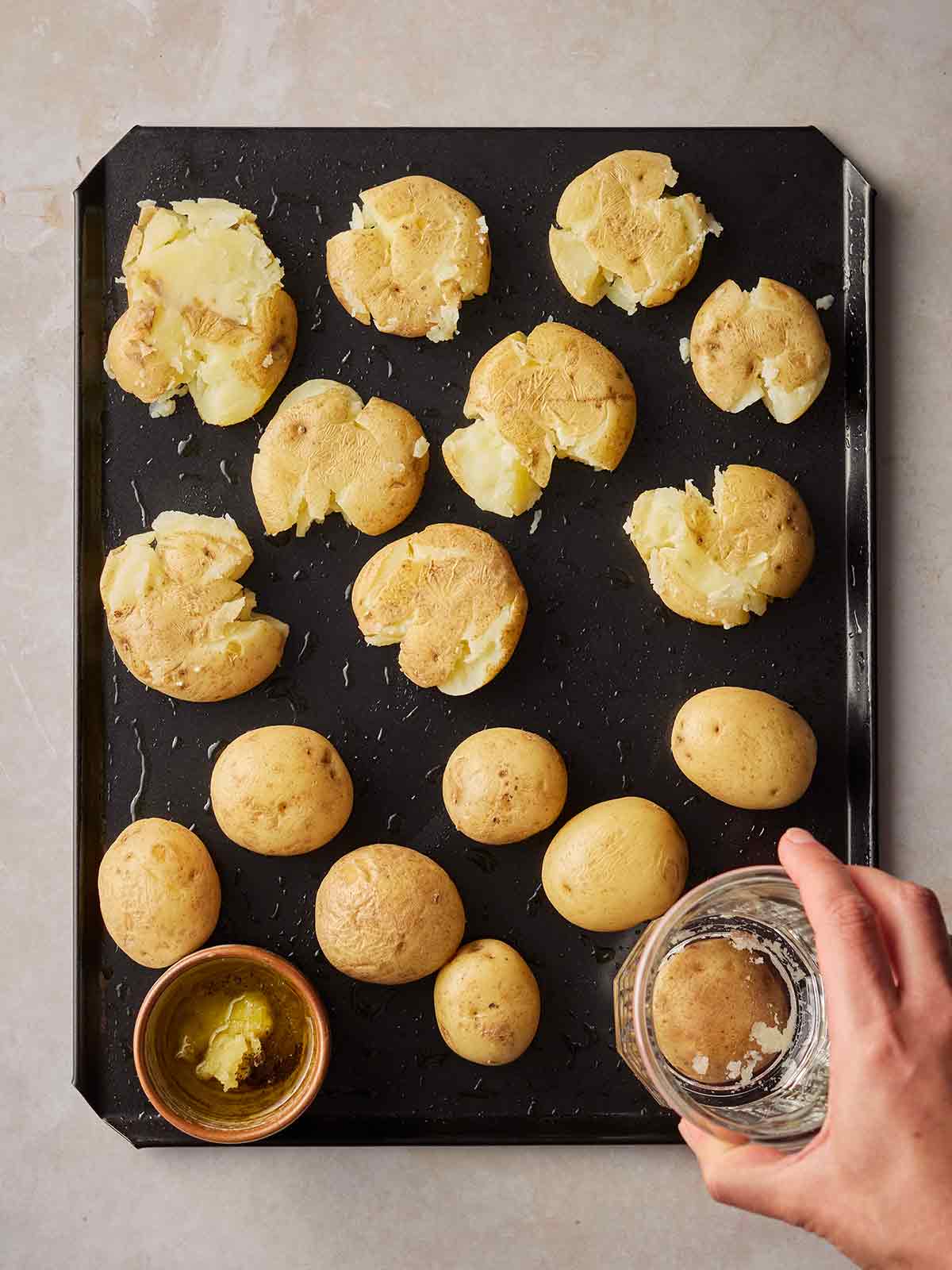 A black baking tray on a white surface, with potatoes being smashed by a hand smashing a glass down onto them.