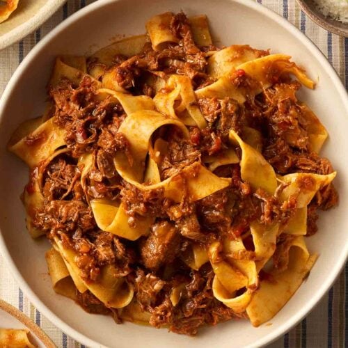 A bowl of beef ragu with pappardelle strips on a table with a stripy tablecloth.