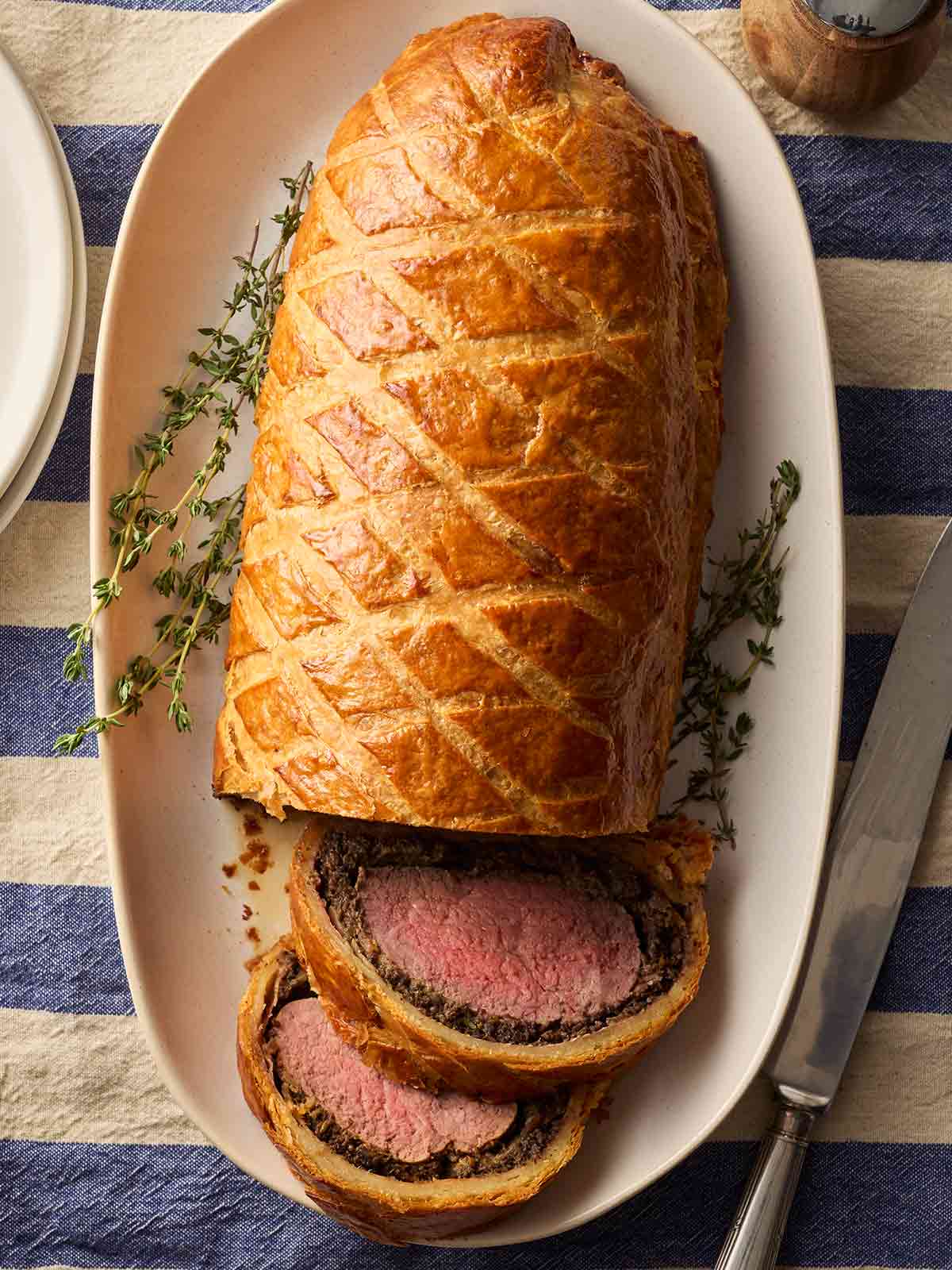 A cooked Beef Wellington with a golden crust laid out on a serving platter with sprigs of thyme, on a blue and white tablecloth, ready to serve.