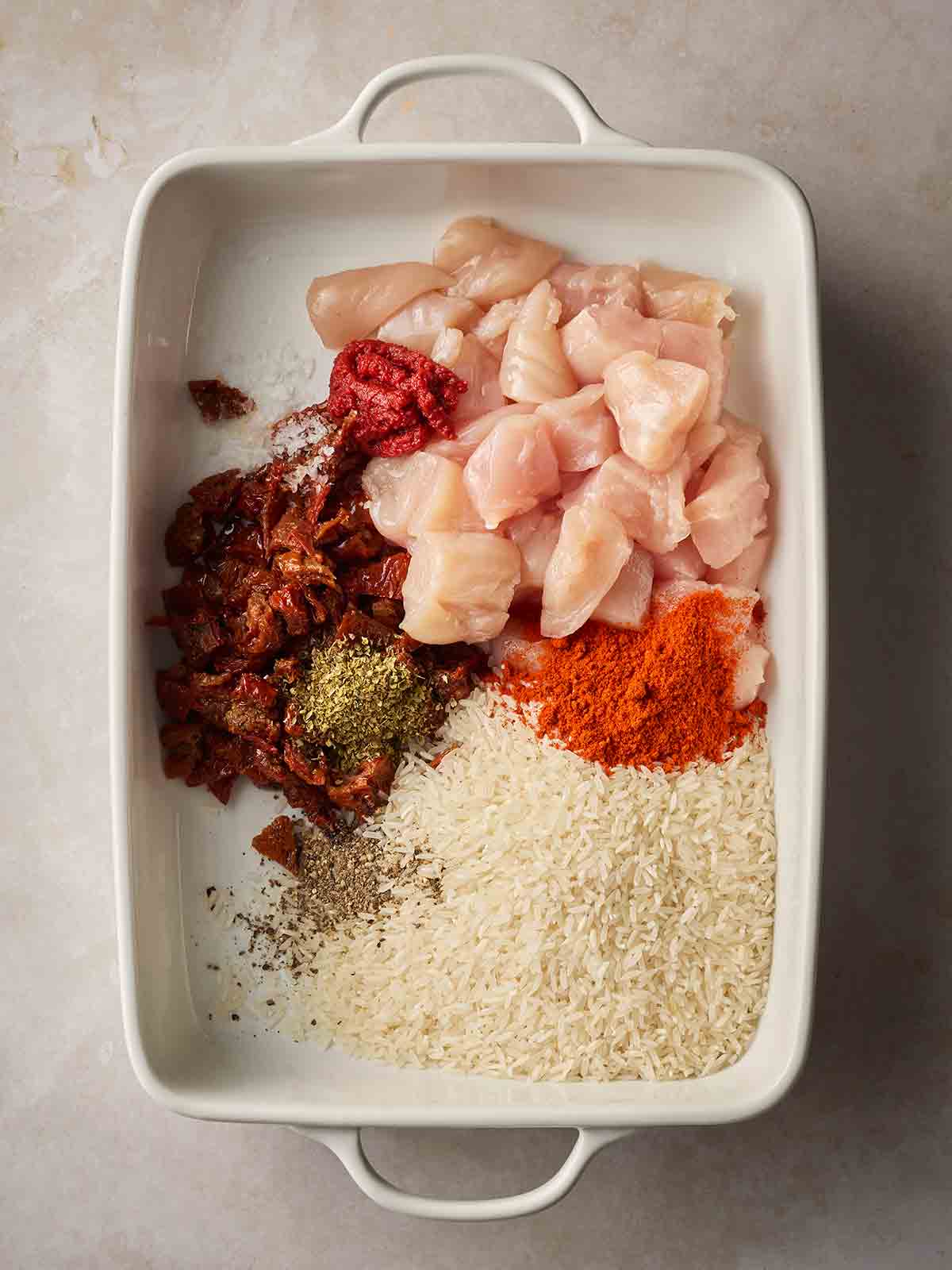 Raw ingredients including chicken cubes, rice, sun-dried tomatoes and herbs, in an oven dish, ready to cook together in the oven.