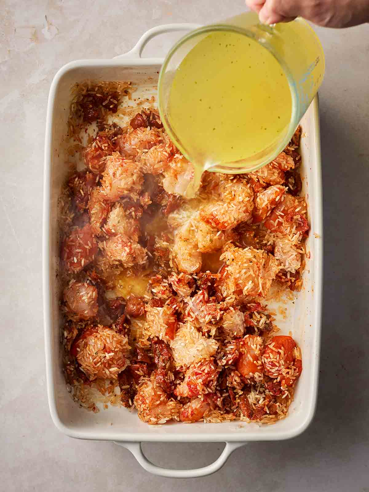 A jug of stock being poured over an oven dish with chicken, tomatoes and rice in.