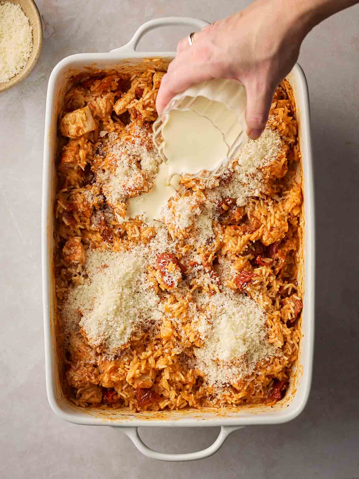 A hand pouring cream into an oven dish of cooked chicken and rice.