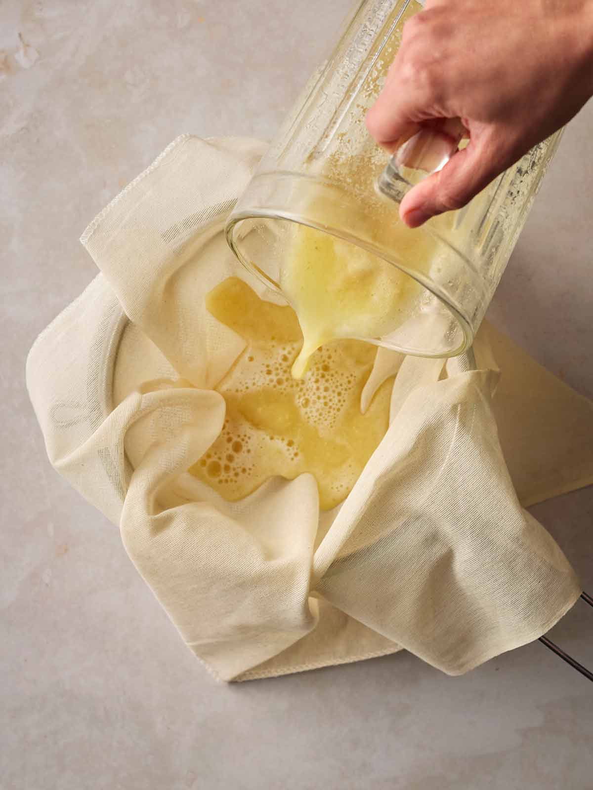 A hand pouring a jug of blended gingers and apple juice through a muslin into a bowl.