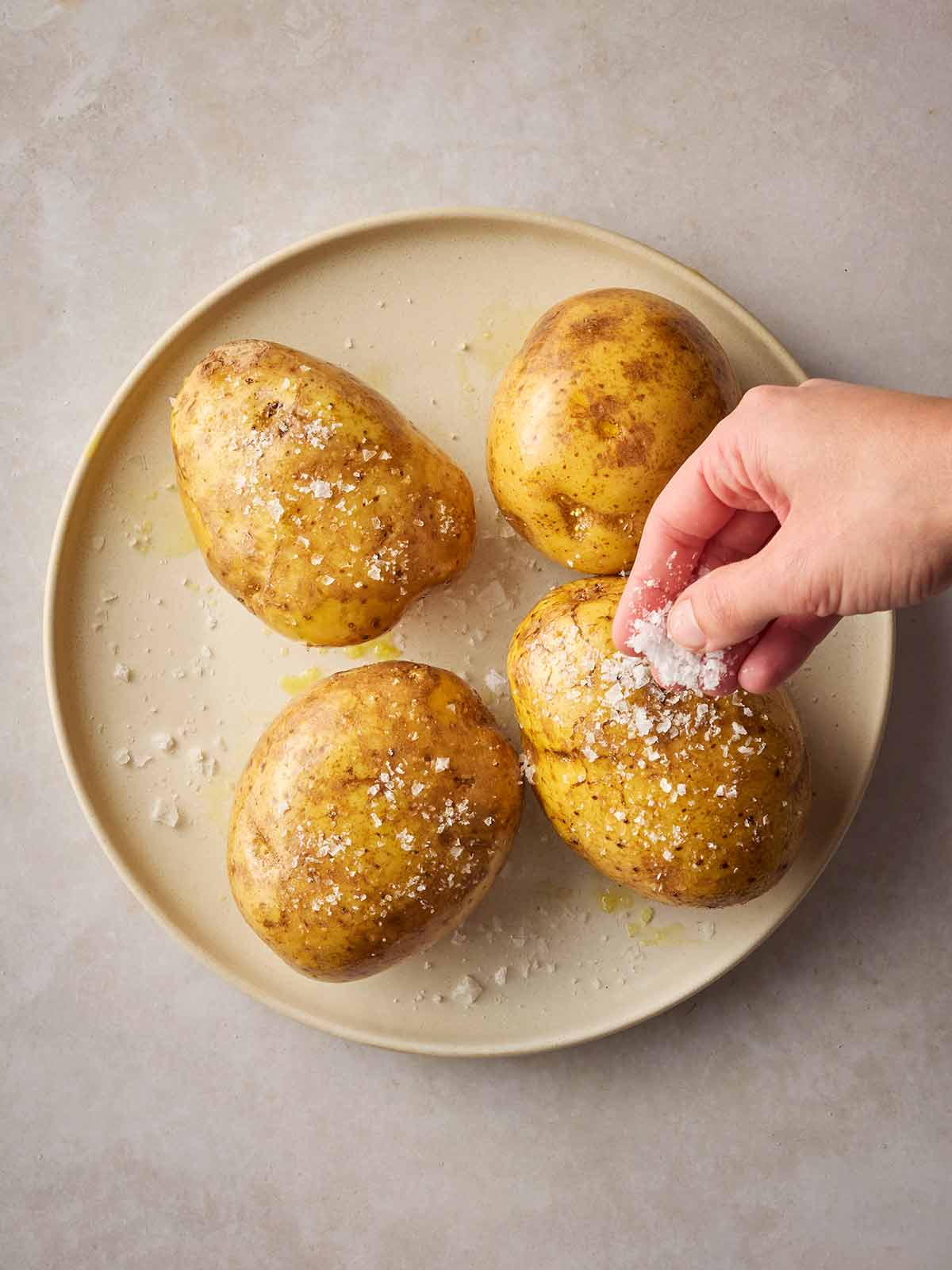 A plate with four oiled white potatoes on, with a hand sprinkling over sea salt.