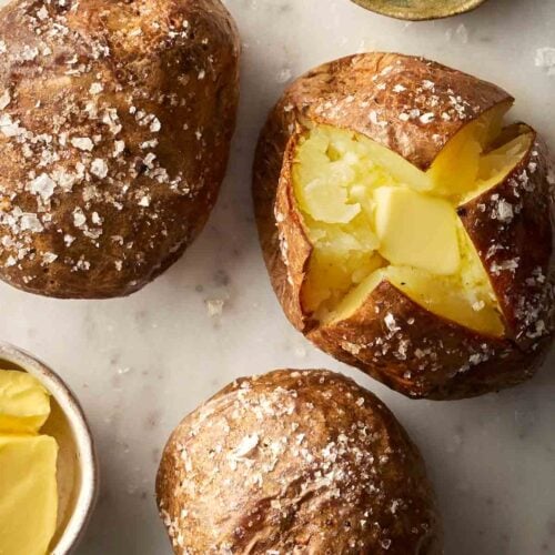 Three cooked baked potatoes on a white surface, with salt and butter to the side.
