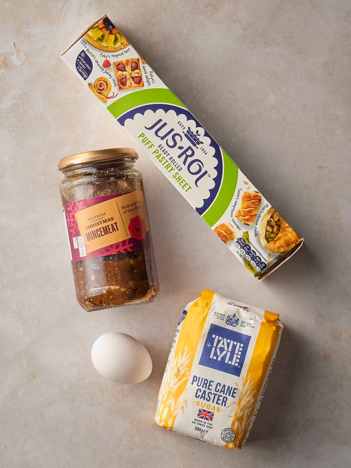 A surface with ingredients on for making mince pies, including puff pastry, caster sugar, an egg and mincemeat in a jar.