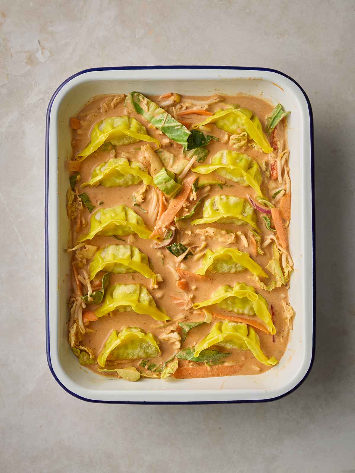 A red thai sauce in an oven dish with dumplings lined up in the pan, with stir fry vegetables around the side, ready to bake.