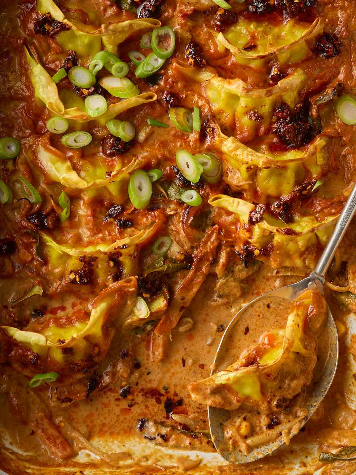 A close up of Chinese dumplings in a red Thai sauce with spring onions over the top and a spoon to serve up.