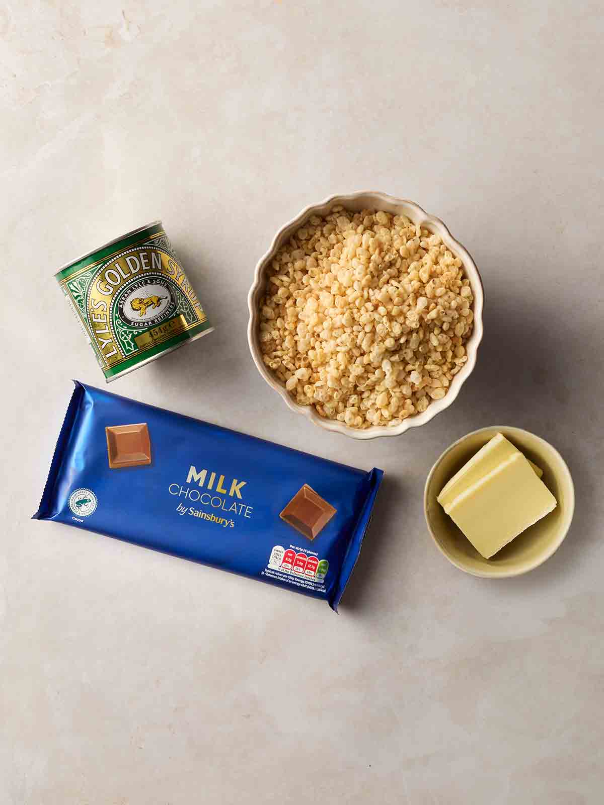 A white surface with ingredients laid on it, including a bar of milk chocolate, a tin of golden syrup, a bowl of Rice Krispies and a bowl of butter.