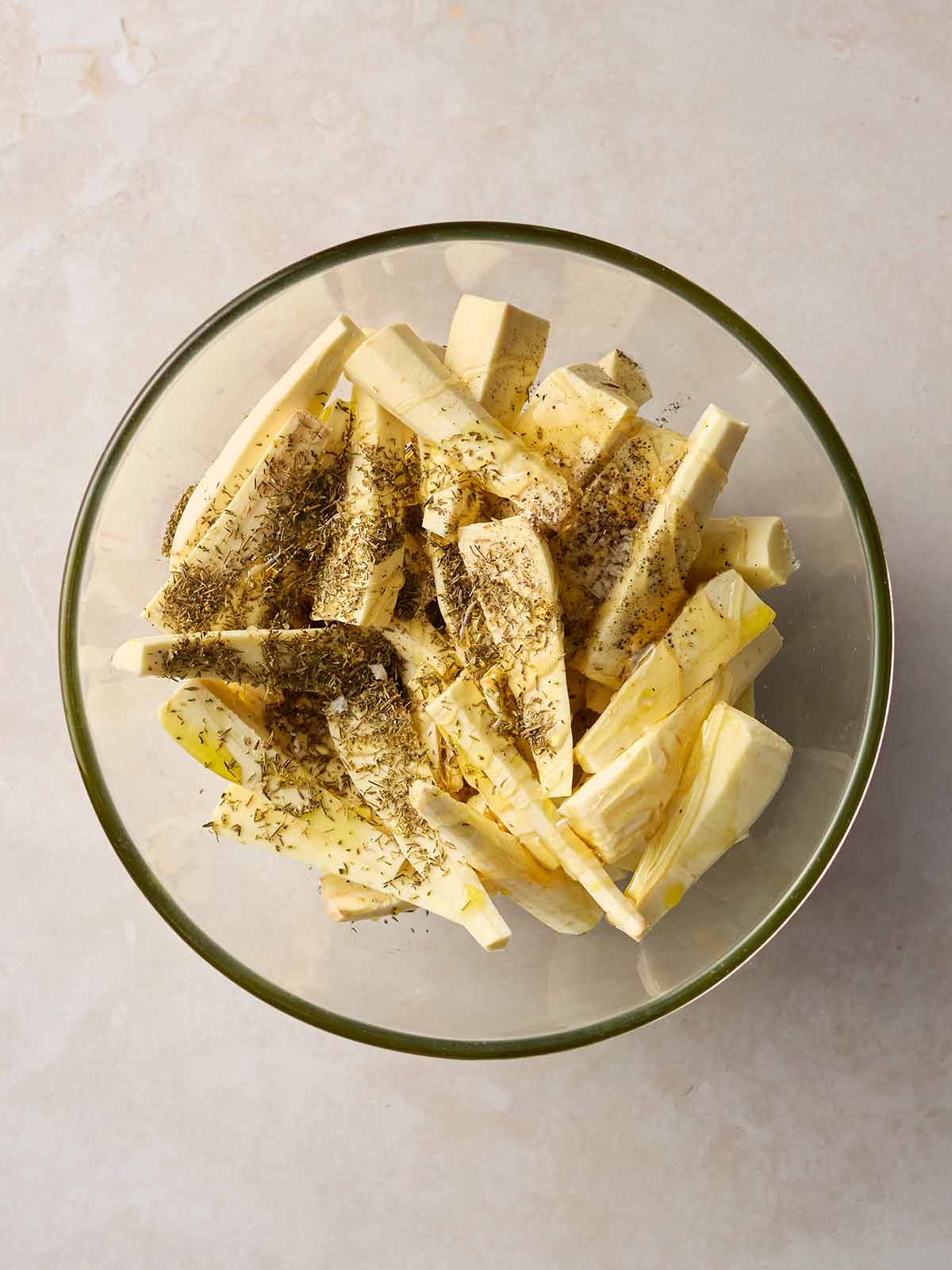 A glass bowl on a white surface, filled with raw chopped parsnips that's covered with dried thyme and honey.
