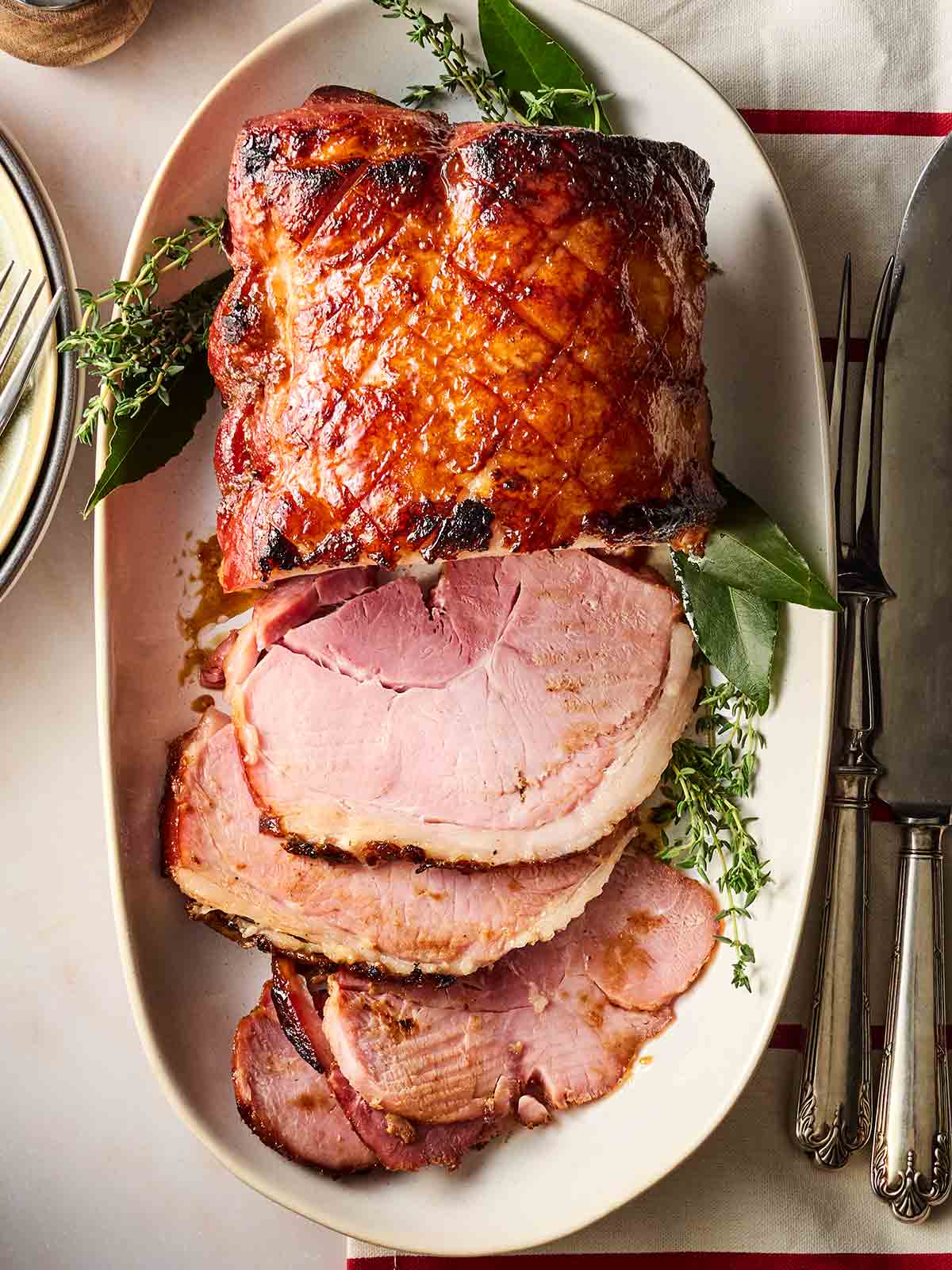 A slow cooker ham on a white platter with sprigs of greenery, half sliced up.