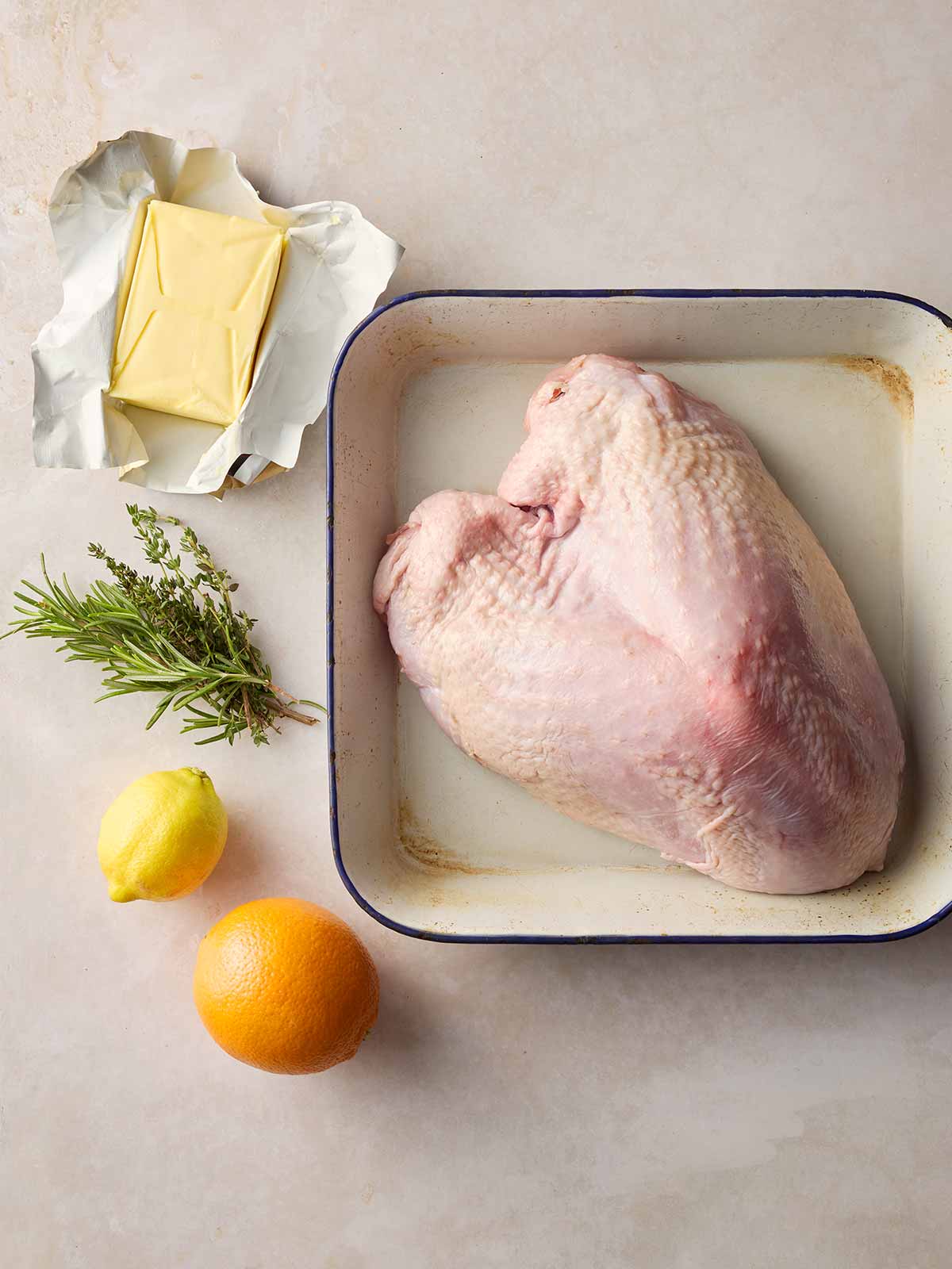 A raw turkey crown in an oven dish on a white counter, surrounded by a block of butter, fresh herbs and an orange and lemon.