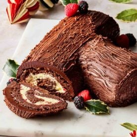 A Christmas yule log on a board, with a slice out to show the insides, and decorated with holly and berries.