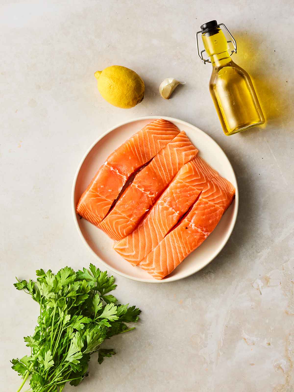 A plate of raw salmon fillets with other ingredients for baking it round the side, including oil, lemon, garlic and parsley.