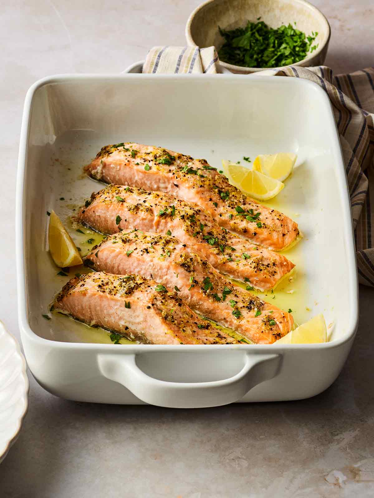 An oven dish with four pieces of baked salmon, with a delicious oil dressing and lemon wedges.