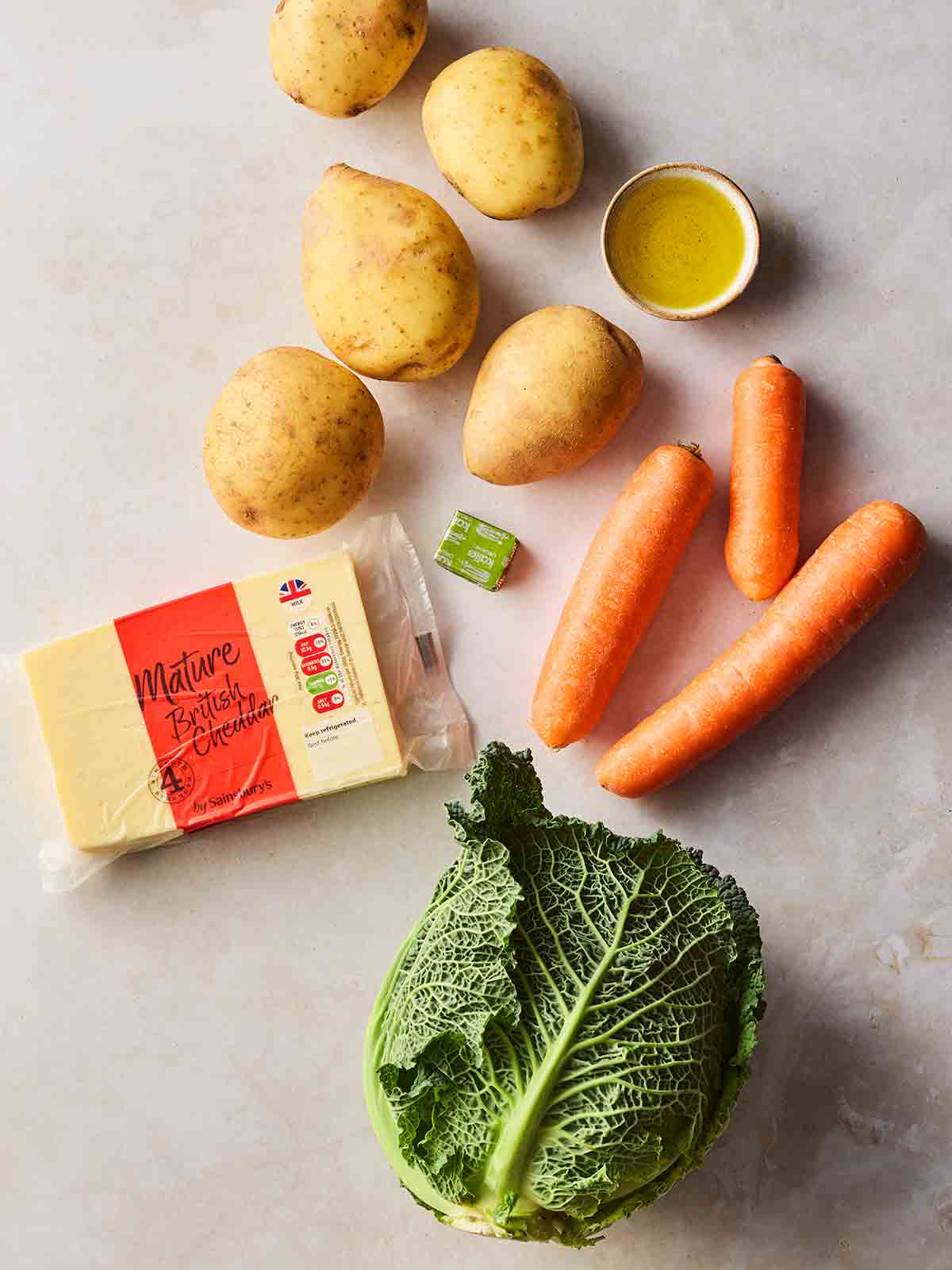 Ingredients laid out on a white counter, including potatoes, oil, carrots, cheese, a stock cube and cabbage.