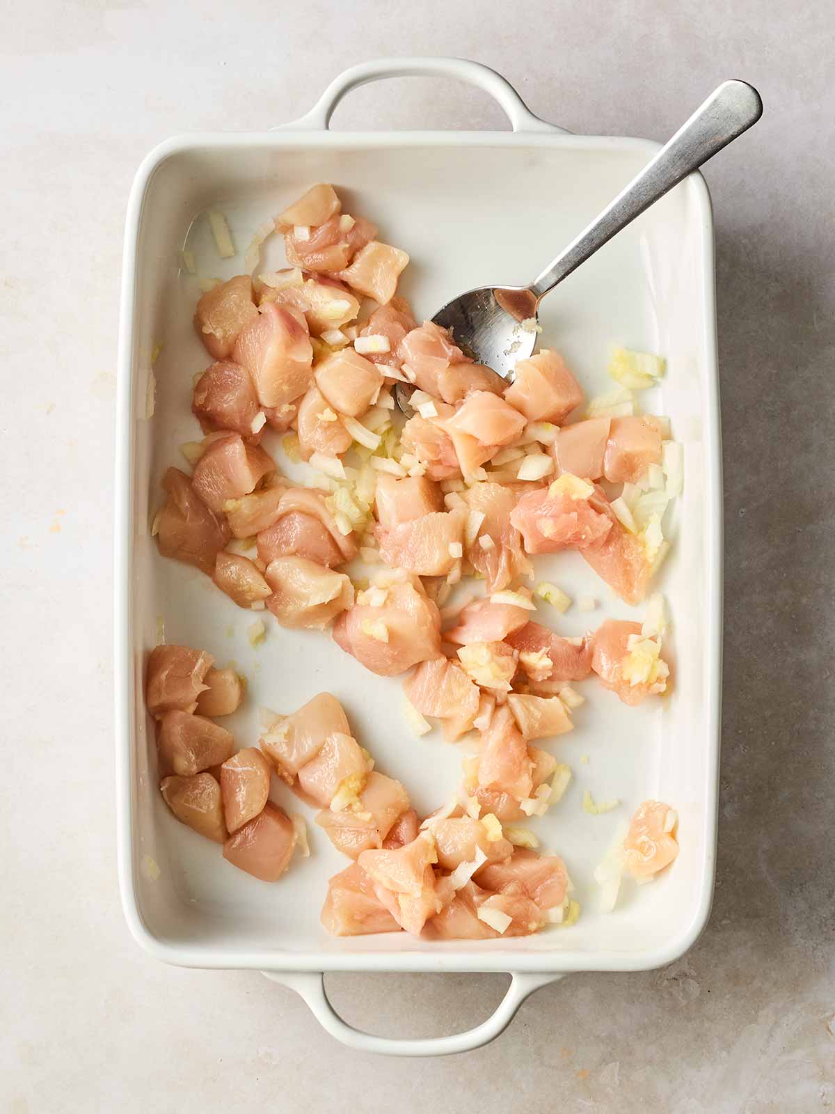 Cubed raw chicken in an oven dish with onion and a spoon.