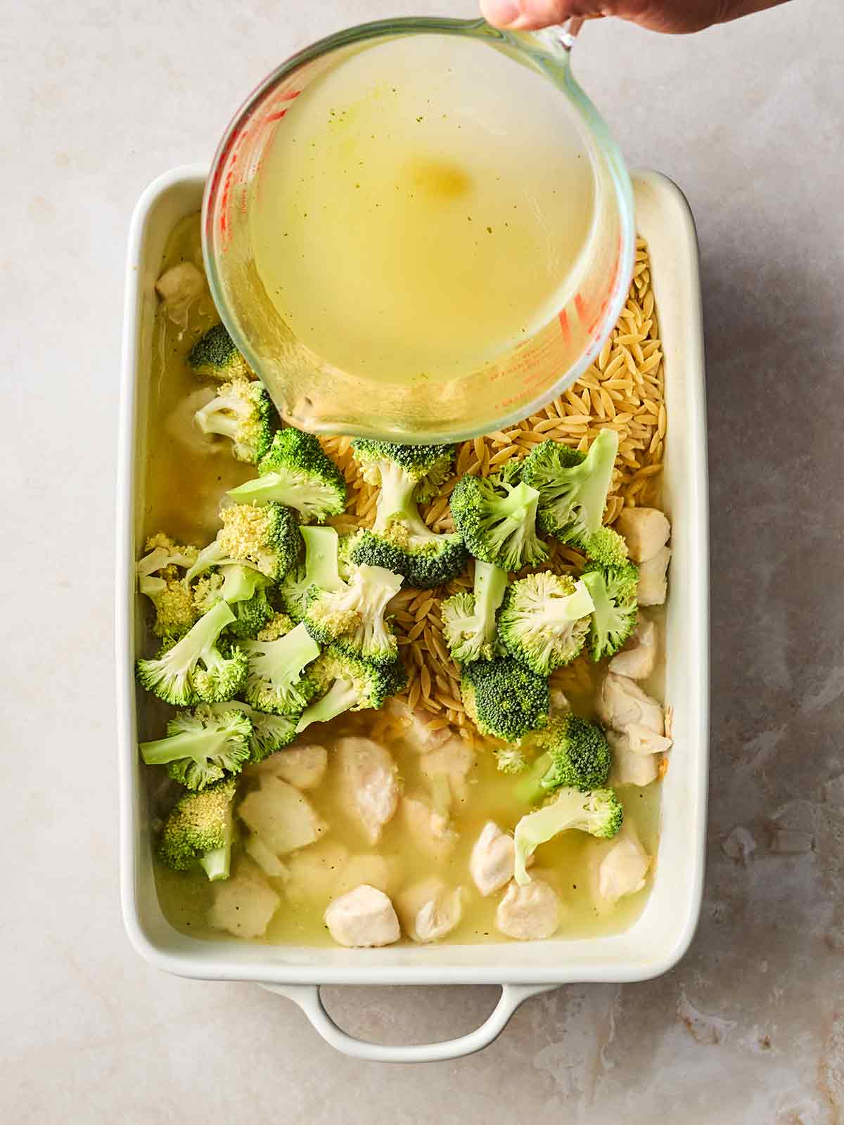 An oven dish with chicken, orzo, broccoli and a hand pouring in chicken stock from a jug.