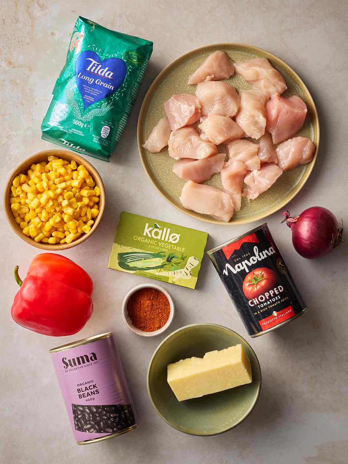 Raw ingredients laid out on a surface ready to make a Burrito Rice dish, including chicken, rice, sweetcorn, peppers, tomatoes, onion, black beans, cheese, stock cubes and paprika.