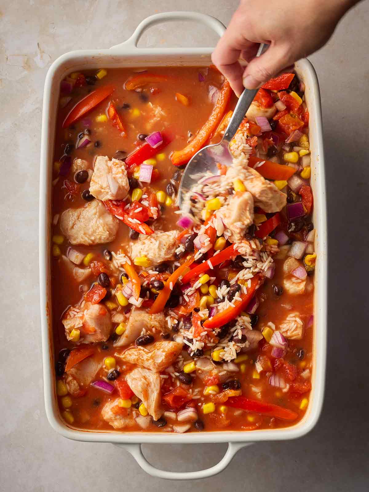 A hand with a spoon stirring an oven dish with half cooked rice and chicken.
