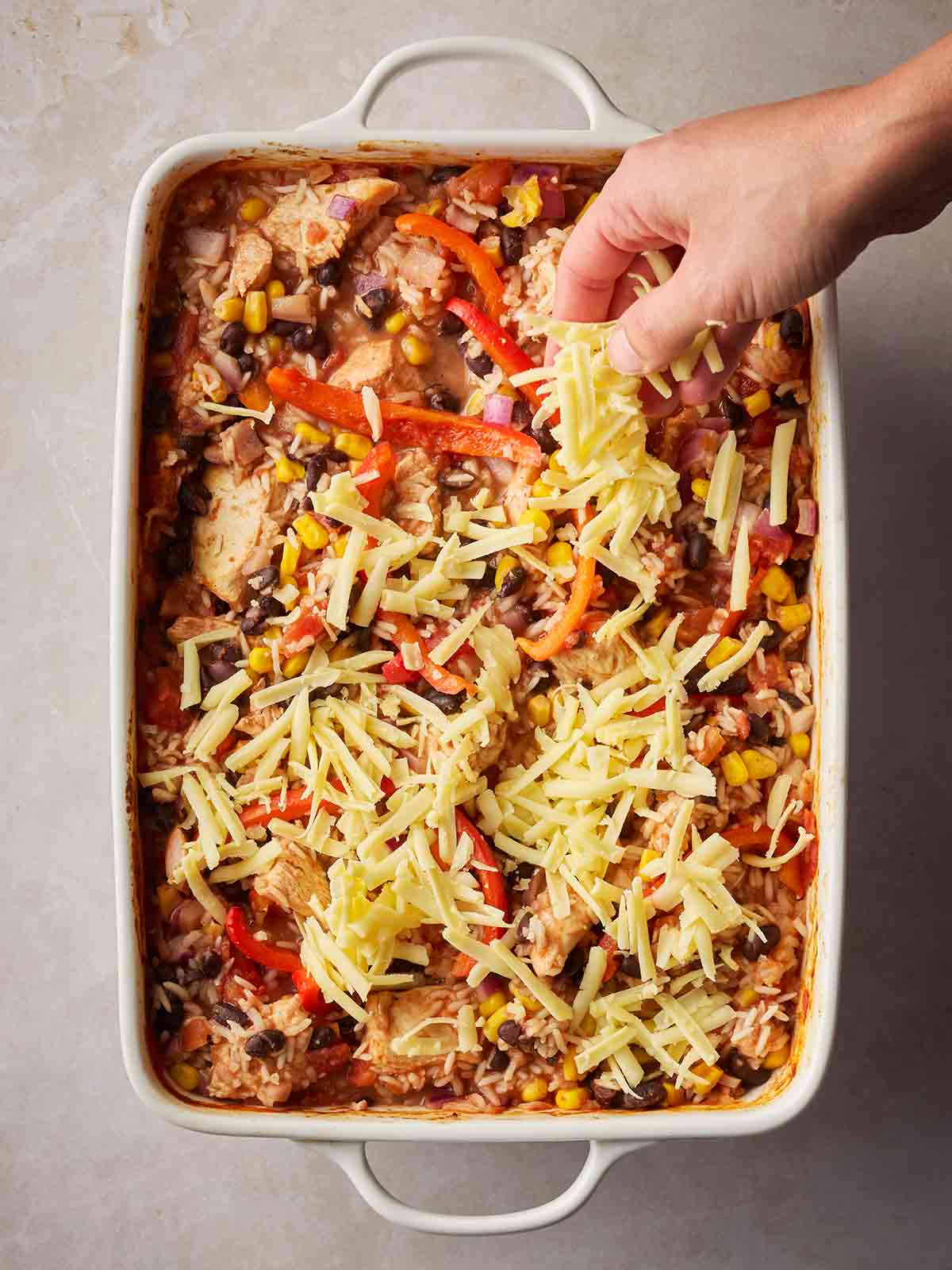 A hand sprinkling grated cheese over the top of an oven dish with chicken and rice burrito style dinner.